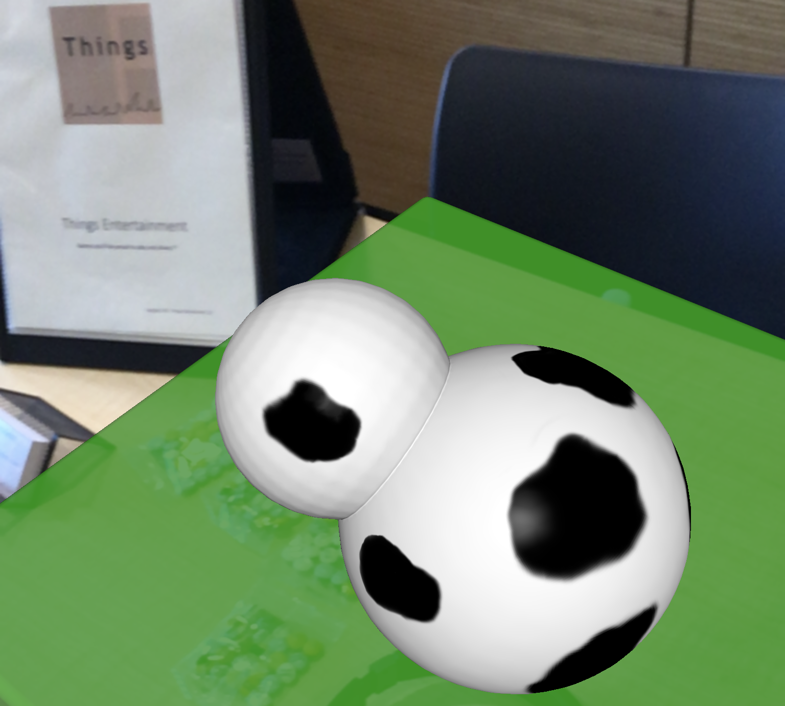 View of a table through an augmented reality application. The table has been augmented with a green field and a demo cow. The green field is semi-transparent, so you can still see other items on the table through the field. The demo cow, though, is opaque.