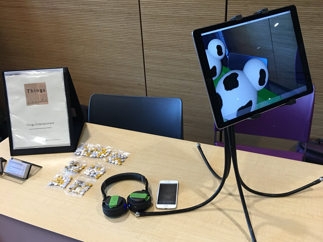 An iPad on a stand is angled down to point at a conference exhibit table. The screen of the iPad shows the table covered with a green cartoon field. On this field there are two really silly 3D demo cows (each cow is just two white spheres combined together with black spots). Also shown: headphones, iPhone, business cards, branded flip book, and branded candies.